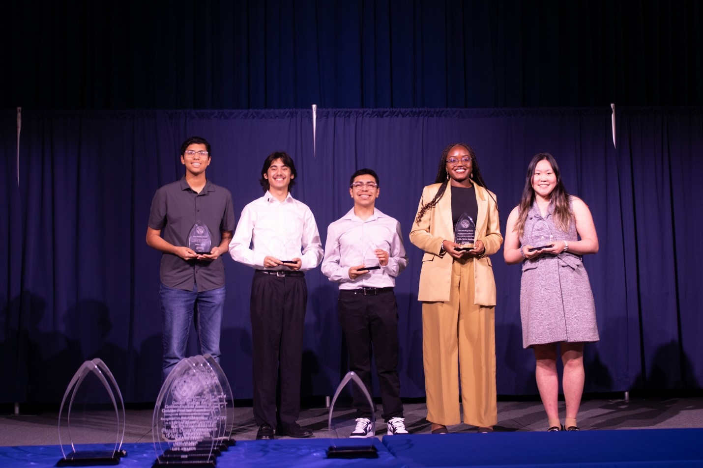 Five students dressed in slacks and button down shirts; one in a dress each holding an award plaque