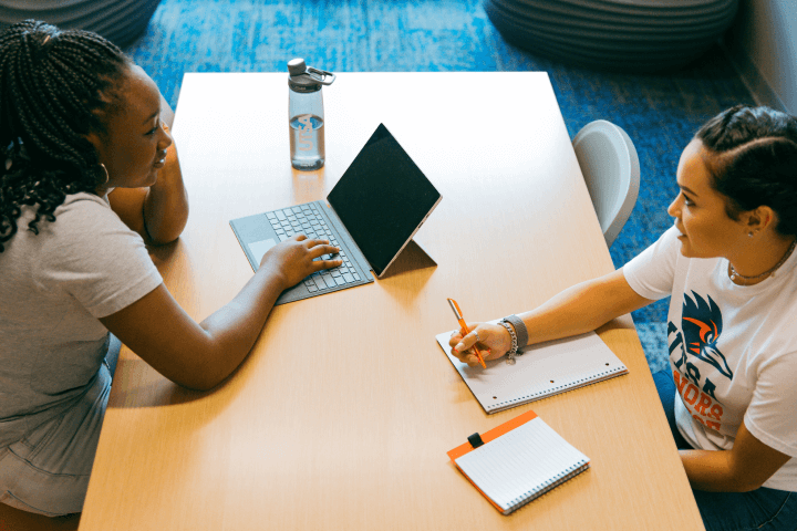 A female student with a laptop and another female student with a notebook sitting at a table