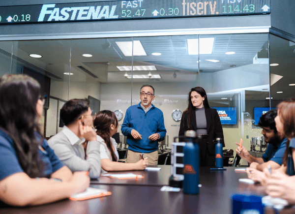 A group meeting in a glass-walled office, led by a man in a blue sweater.