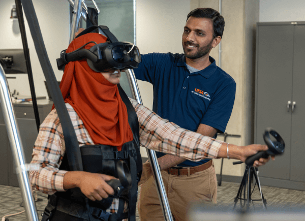 A woman wears a VR headset and harness in a lab setting, holding controllers.