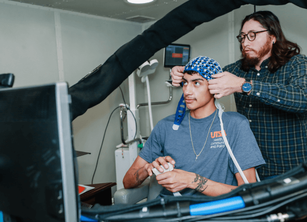 Man adjusts a blue mesh cap on a seated person's head in a lab setting.