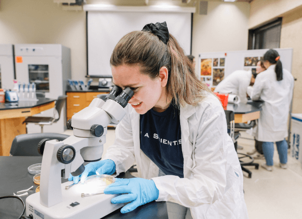 A young scientist in a lab coat and gloves looks through a microscope in a bright laboratory.