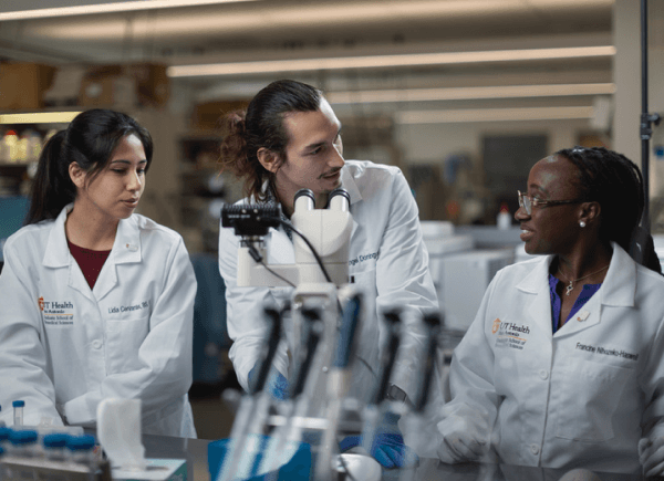 Three researchers in lab coats are discussing a project around a microscope in a laboratory.