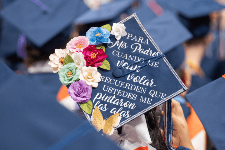 Graduation cap decorated with colorful flowers, a butterfly, and Spanish text honoring parents.