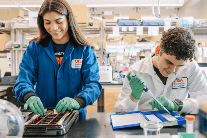 Two scientists in a lab, wearing UTSA-branded lab coats and gloves.