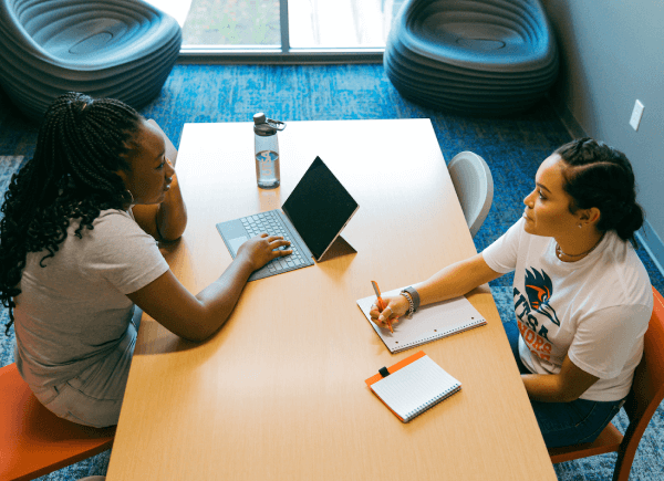 Two women sit across a wooden table in an office setting, engaged in conversation.