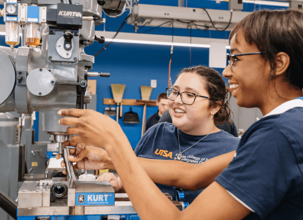 Two students in a workshop operate a milling machine, smiling and engaged.