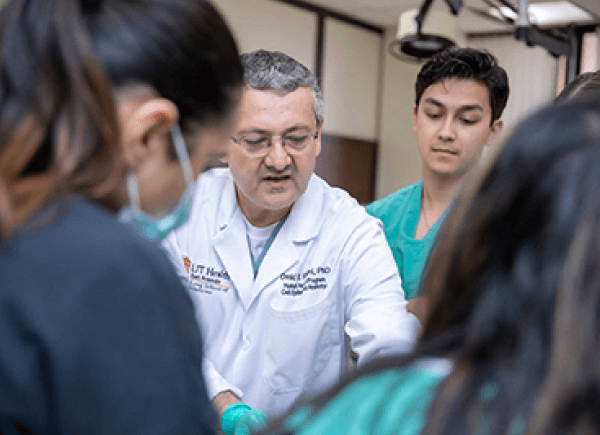 A male doctor in a white coat, surrounded by medical students, instructs a procedure.