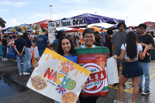 two students promoting nachos with a poster and dressed as salsa, in front of row of booths